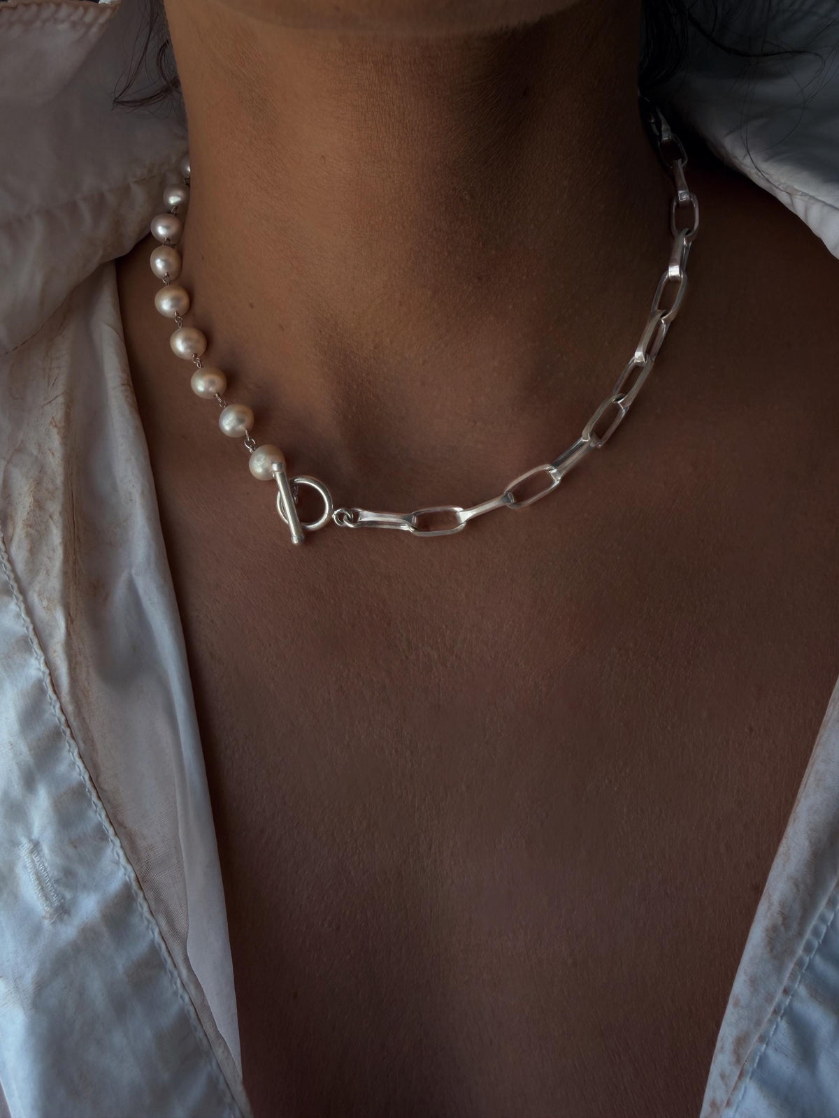May include: A necklace featuring a string of white pearls on one side and a silver-toned metal chain with elongated oval links on the other. The two sections are connected by a silver-toned toggle clasp positioned at the front of the neck.