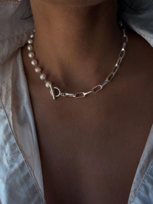 May include: A necklace featuring a string of white pearls on one side and a silver-toned metal chain with elongated oval links on the other. The two sections are connected by a silver-toned toggle clasp positioned at the front of the neck.