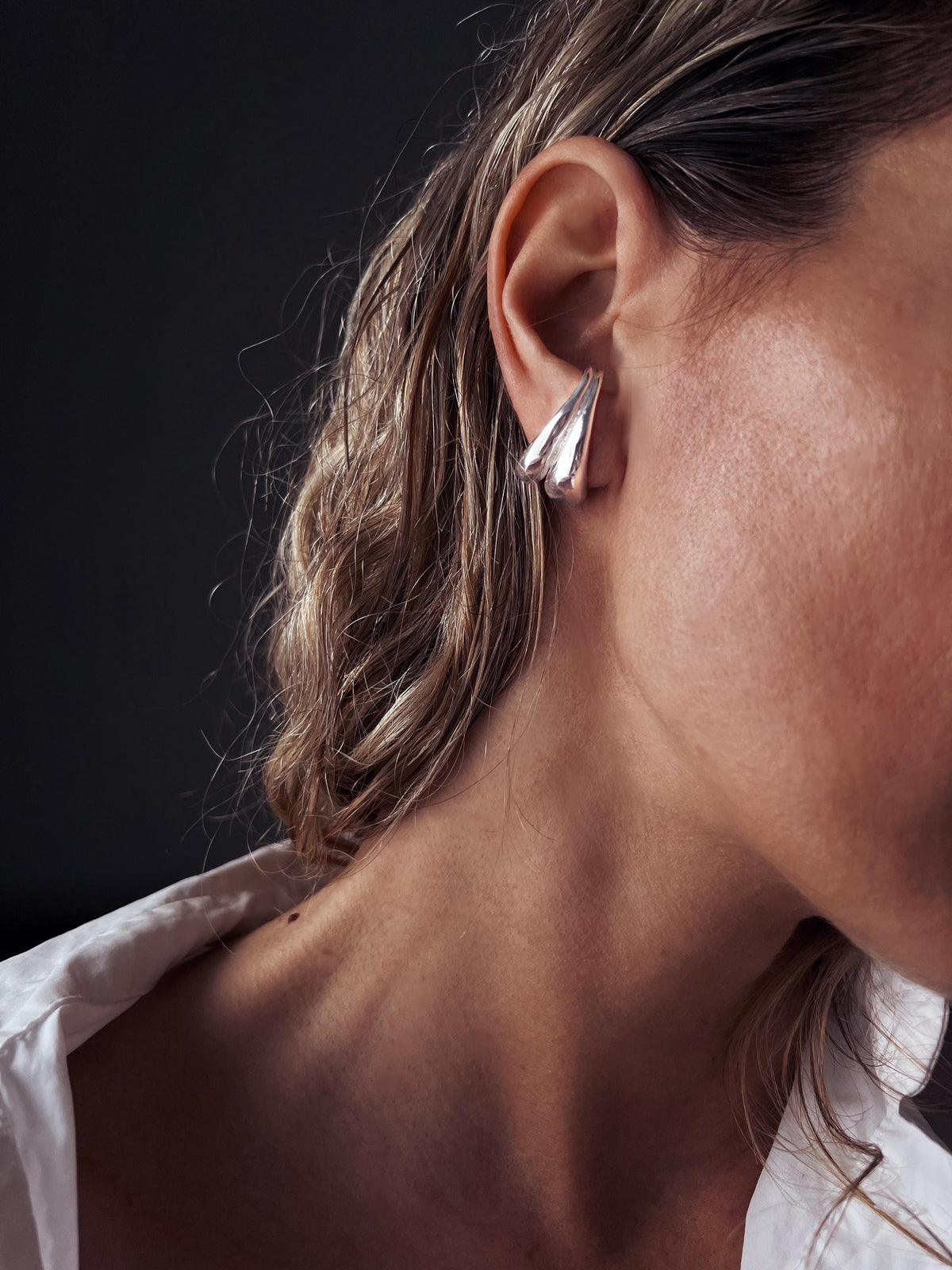 May include: Silver-toned earrings with a layered, triangular design. The earrings are worn on the ear, with a dark background. The earrings have a smooth, reflective surface, and the design is geometric and modern.