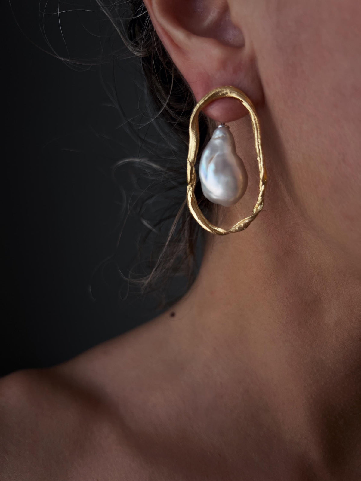 May include: A close-up of a gold-colored earring with a large, irregularly shaped white pearl. The earring has a textured, oval-shaped frame. The background is a dark, neutral tone.