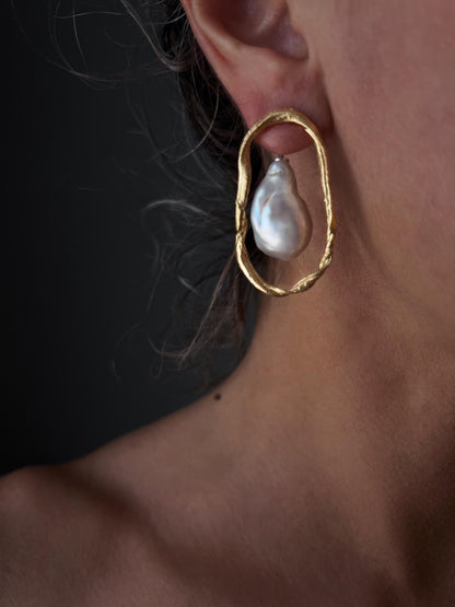 May include: A close-up of a gold-colored earring with a large, irregularly shaped white pearl. The earring has a textured, oval-shaped frame. The background is a dark, neutral tone.