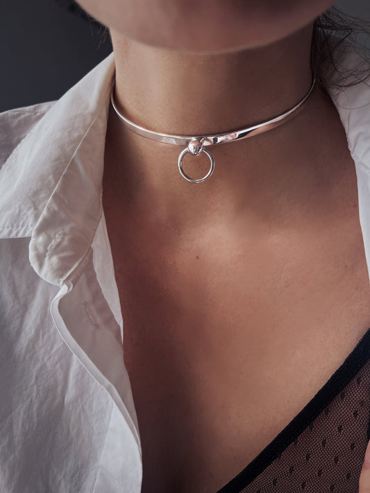May include: A silver choker necklace with a round ring pendant. The necklace is made of a thick, flat band of metal.