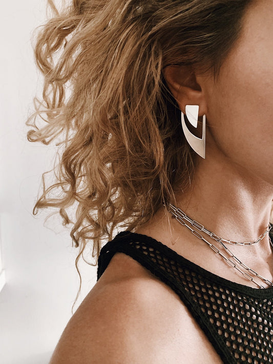 May include: A woman with curly blonde hair wearing a black mesh tank top and silver geometric earrings.