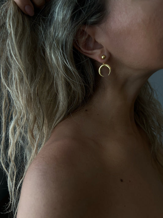 May include: Gold-colored crescent moon earrings. The earrings have a small, round stud at the top and a curved, open-ended crescent shape hanging below. The jewelry is worn on a person's ear.
