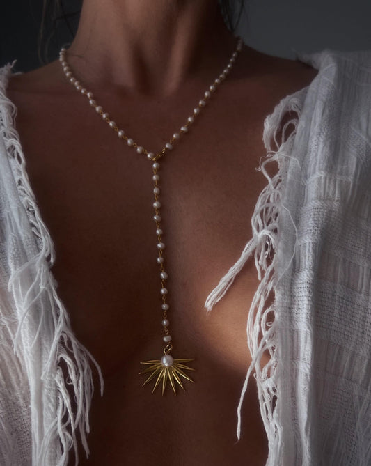 May include: A delicate pearl necklace with a gold sunburst pendant. The necklace features small, round pearls strung together with a gold chain. The pendant is a gold sunburst design with a single pearl at its center. The necklace is worn over a white garment.