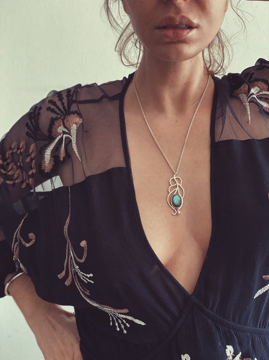 May include: A silver necklace with a turquoise gemstone pendant in the shape of a leaf. The necklace is worn by a person with a black sheer top with floral embroidery.